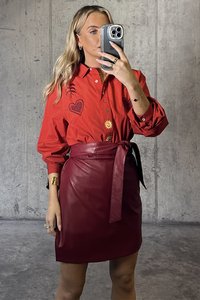 Punainen ylisuuri paita, jossa on brodeerattuja sydämiä ja aurinko, yhdistettynä korkeavyötäröiseen viininpunaiseen nahkahameeseen, jossa on sidottu vyöyksitys.