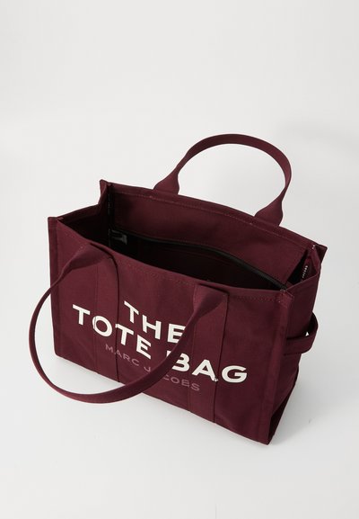 Borsa a tracolla in tela bordeaux con manici doppi, caratterizzata da un design grafico bianco che riporta la scritta "THE TOTE BAG MARC JACOBS." Apertura superiore con tasca interna.