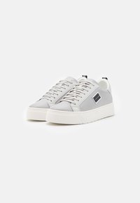 Baskets gris texturé avec une semelle en caoutchouc blanche, présentant des lacets plats et un petit logo noir sur le côté. Design minimaliste.