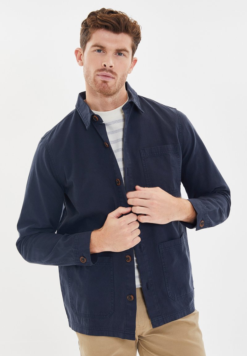 Giacca button-up blu navy in cotone, con colletto, due tasche anteriori e bottoni marroni, indossata sopra una maglietta a righe.