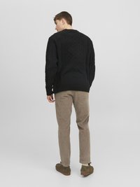 Maglione lavorato a maglia nera con motivo a treccia, indossato con pantaloni di velluto a coste marrone chiaro e scarpe verdi. Modello girato di spalle rispetto alla fotocamera.