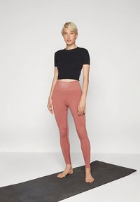 Črna skrajšana majica v kombinaciji z roza legicami z visokim pasom, ki imajo na pasu sončni vzorec, nosi oseba, ki stoji na blazini.