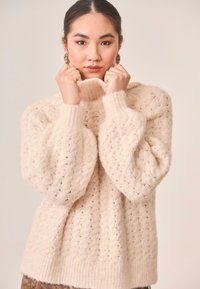 Suéter beige claro de punto con cuello alto, patrón de diamantes texturizado y mangas abullonadas, hecho de un material suave y esponjoso.