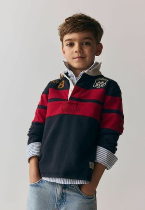 Chlapec s krátkými hnědými vlasy, oblečený v námořnicky modré a červené proužkované ragbyové košili přes modrou proužkovanou košili na knoflíky, ruce v kapse světle modrých džínů, neutrální pozadí.