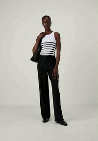 Λευκό αμάνικο μπλουζάκι με μαύρες ρίγες, μαύρο παντελόνι σε φαρδιά γραμμή και μαύρη υφασμάτινη τσάντα. Το μοντέλο στηρίζεται όρθιο, φορώντας μαύρα παπούτσια.