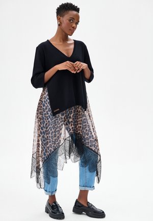 Kobieta stojąca, ubrana w czarny sweter z dekoltem w serek, przezroczystą asymetryczną spódnicę w panterkę nałożoną na podwinięte jeansy oraz czarne mokasyny na białym tle.