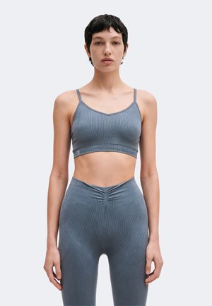 Mujer de pelo corto y oscuro de pie de frente, lleva un top corto gris ajustado con tirantes finos y unos leggings de talle alto a juego.