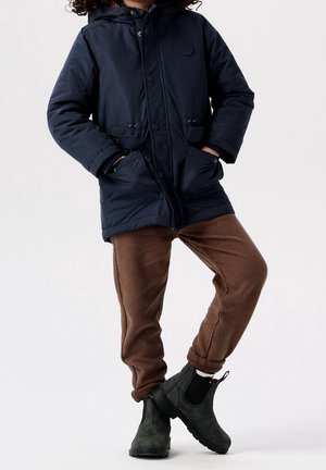 Enfant portant une veste à capuche bleu marine, un pantalon marron et des bottines vert foncé, debout les mains dans les poches.