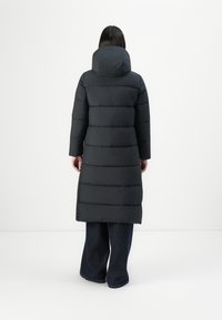 Vrouw met haar rug naar de camera gekeerd, gekleed in een lange zwarte gewatteerde jas met een capuchon en wijdvallende donkerblauwe broek, tegen een eenvoudige witte achtergrond.