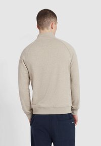 Beige sweatshirt met een hoge kraag, ribgebreide manchetten en een ribgebreide zoom. Heeft een zachte textuur en raglanmouwen. Ggedragen met donkerblauwe broek.