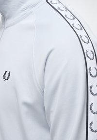 Gros plan d'une manche de veste blanche avec un col côtelé et des logos de couronne de laurier noirs le long de la bande d'épaule et de la poitrine.