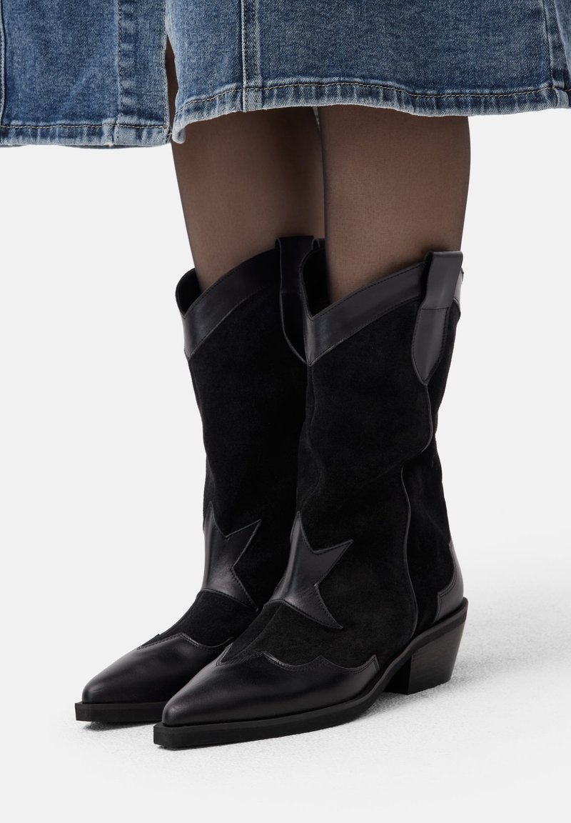 Botoși cowboy din suede negru cu accente de piele, vârful ascuțit și toc mic pătrat. Prezintă detalii în formă de stea pe părți.