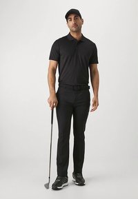 Svart polo skjorta och byxor bärs med en svart keps och golfskor, håller en golfklubba. Outfiten är sportig och åtsittande.
