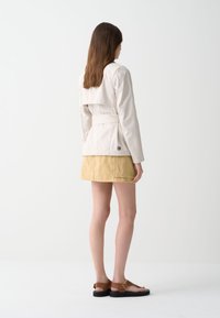 Trench-coat beige clair avec une taille cintrée, présentant un détail de rabat au dos, associé à une jupe jaune moutarde et des sandales marron.