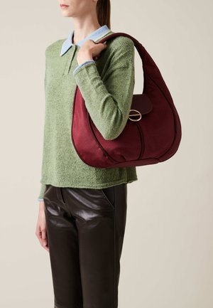 Weinrot strukturiertes Handtäschchen mit gebogener Form, einem Klappenverschluss und einem goldenen Hardware-Detail, gehalten von einer Person in einem grünen Pullover und Lederhosen.