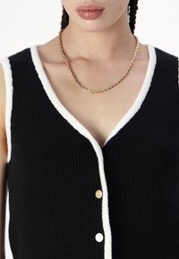 Žena oblečená v černé bezrukávové pletené vestě s bílým lemem a zlatými knoflíky, a se zkrouceným zlatým řetízkem.