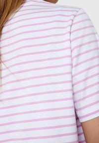 Osoba v bílém krátkém rukávu s horizontálními růžovými pruhy, ukazující horní část zad a pravé rameno.