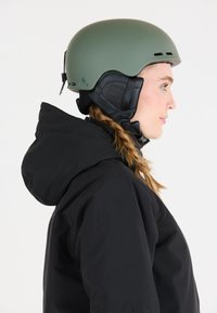 Groene skihand die een gladde, matte afwerking heeft, verstelbare kinband en ventilatieopeningen, gedragen met een zwarte hoodie.
