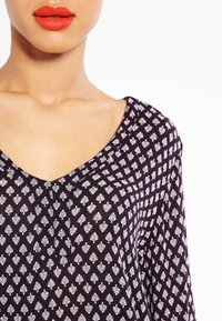 Schwarzer Stoff mit einem weißen Blattmuster; V-Ausschnitt-Design, das Schlüsselbein und Schultern freilegt, glatte Textur im Stoffdetail sichtbar.
