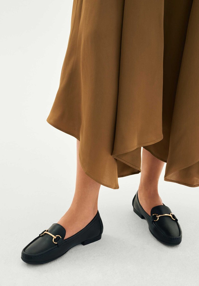 Picioare într-o fustă maro, încălțate cu mocasini negri din piele, cu detaliu auriu în formă de potcoavă, pe un fundal neutru.