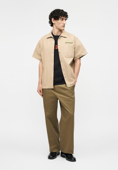 Άνδρας που φοράει μπεζ πουκάμισο με κοντό μανίκι και κουμπιά πάνω από μαύρο μπλουζάκι με στάμπα, χακί φαρδιά παντελόνια και μαύρα δερμάτινα παπούτσια, κοιτάζοντας πλάγια.