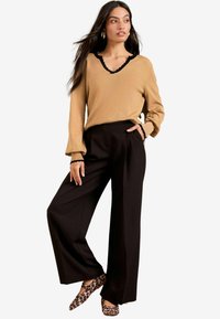 Pull tan avec encolure à volants noirs, associé à un pantalon large noir et des ballerines à imprimé léopard. Tenue élégante, moderne et polyvalente.