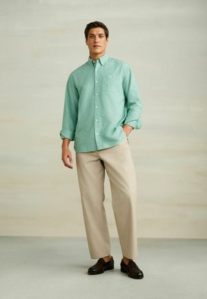 Junger Mann, der ein mintgrünes Hemd mit Knöpfen, beige Hosen und dunkelbraune Loafers trägt, vor einem hellen neutralen Hintergrund steht.