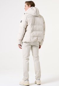 Pufferjack in lichtbeige met een capuchon, voorzien van een gewatteerde structuur en een logo patch op de linker mouw. Gecombineerd met lichtgekleurde broeken en schoenen.