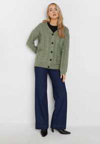 Olivgrön stickad cardigan med kabelmönster, V-ringning och svarta knappar. Kombinerad med mörka vida jeans och svarta klackskor.