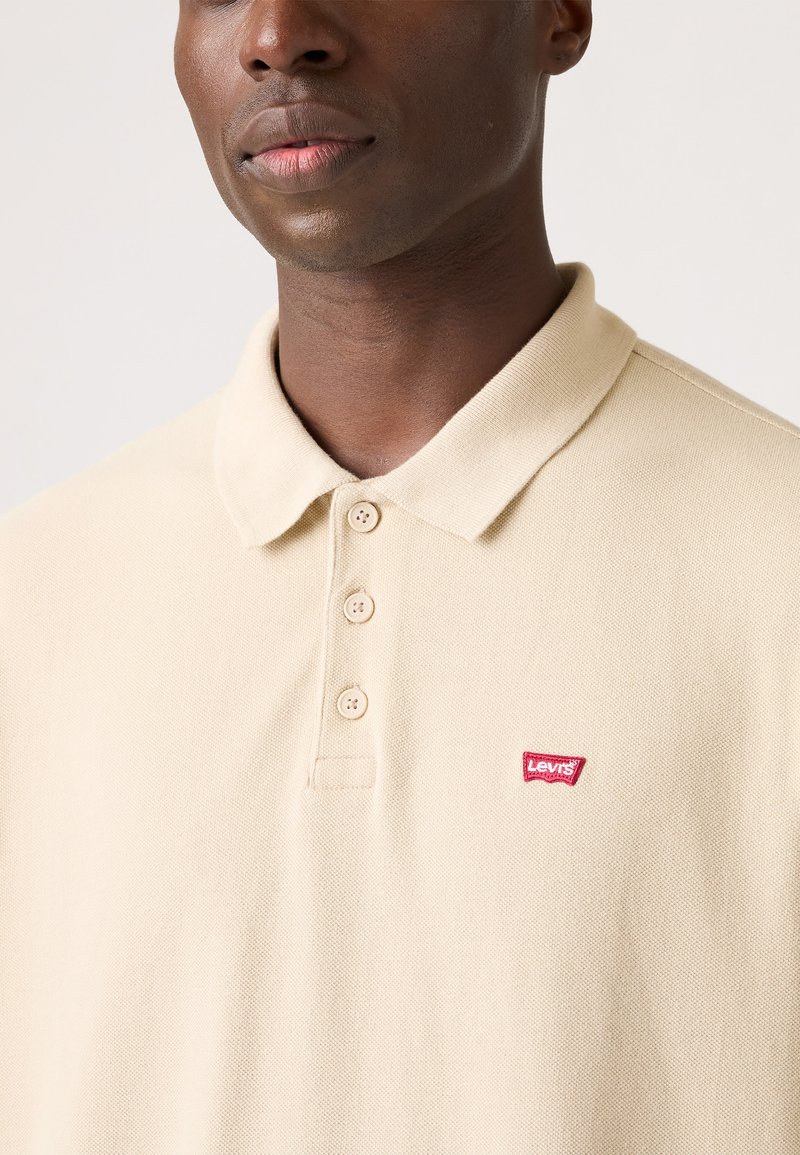 Polo beige con tejido texturizado, que presenta un cuello tipo polo y tres botones. El logo de Levi's se muestra en rojo en el lado izquierdo del pecho.