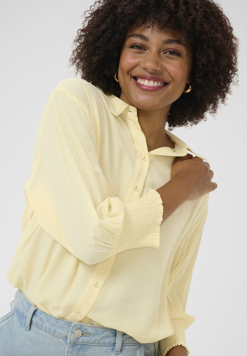 Camicia gialla con bottone, dal fit rilassato, caratterizzata da un colletto, maniche smock testurizzate, abbinata a jeans chiari a vita alta.