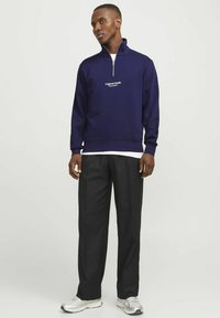 Sudadera navy de medio cremallera con un logo en el frente, combinada con pantalones negros y zapatillas plateadas. La tela parece suave y tiene un ajuste casual.