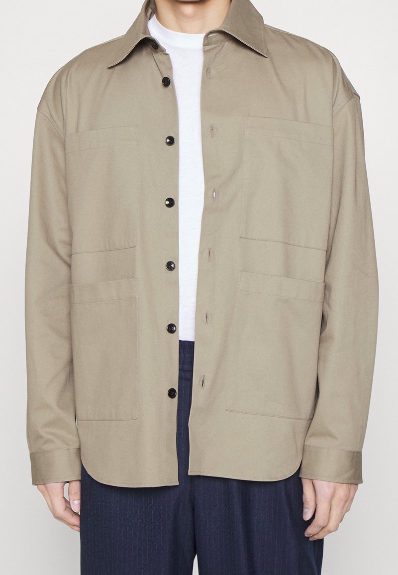 Khaki košile s dlouhým rukávem, zapínáním na knoflíky, dvěma náprsními kapsami a klasickým límečkem. Tkanina je hladká, s uvolněným střihem a rovnými lemy.