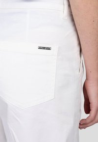 Pantalon en denim blanc présentant une poche arrière avec un design rectangulaire cousu et une petite étiquette de marque métallique. Texture lisse et coupe ajustée.