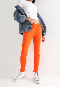 Henkilö, joka wearing kirkkaan oranssit verkkareita, joissa on valkoiset raidat, valkoiset korkeavartiset tennarit, valkoinen lyhyt toppi ja farkkutakki heitetty hartioille.