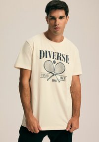 Kremowa bawełniana koszulka z krótkim rękawem, prezentująca niebieski nadruk graficzny z rakietami tenisowymi i napisem "DIVERSE TENNIS CLUB 1993". Luźny krój.