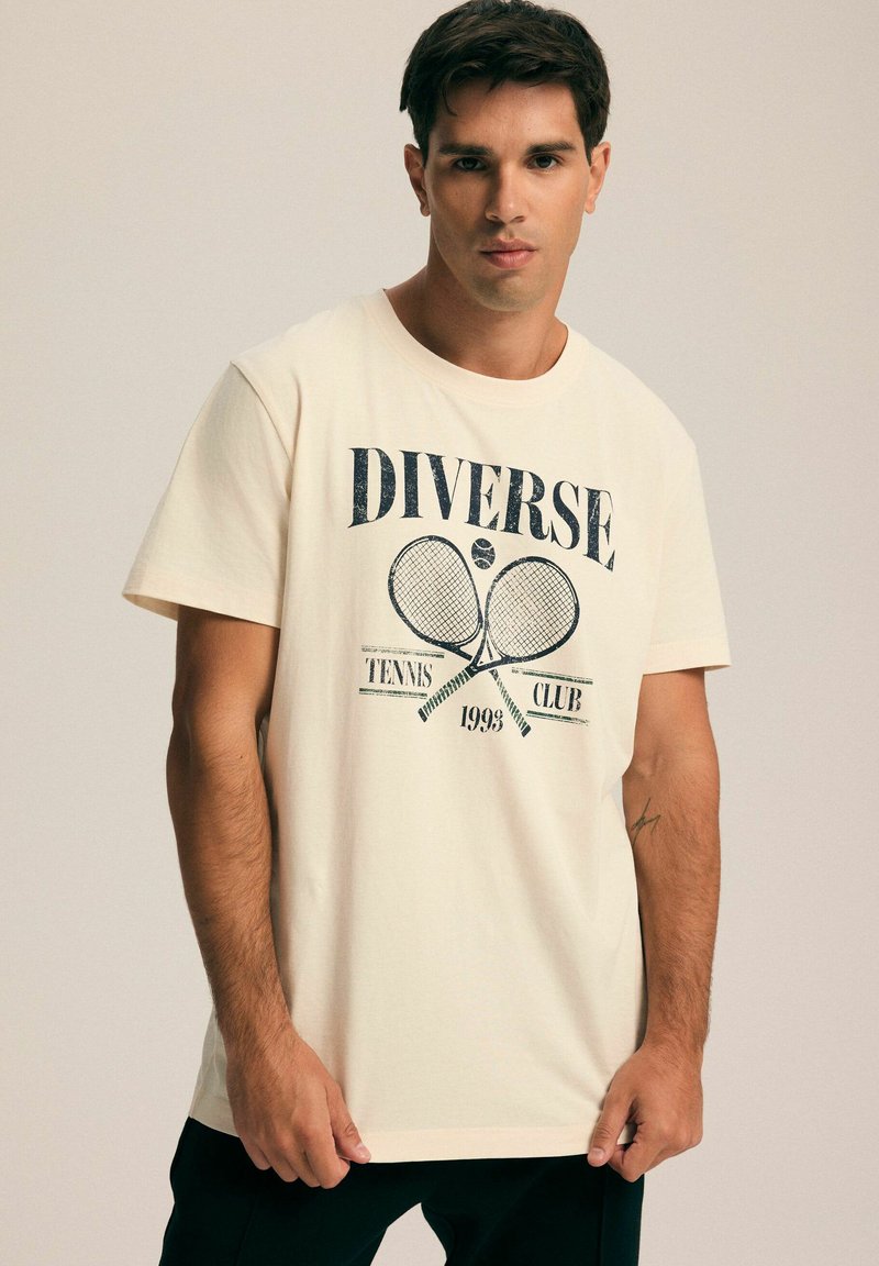 Kremowa bawełniana koszulka z krótkim rękawem, prezentująca niebieski nadruk graficzny z rakietami tenisowymi i napisem "DIVERSE TENNIS CLUB 1993". Luźny krój.