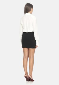Blusa bianca con maniche lunghe e gonna mini nera con dettagli in bottone dorato sul lato. Modella che indossa scarpe con tacco bordeaux.
