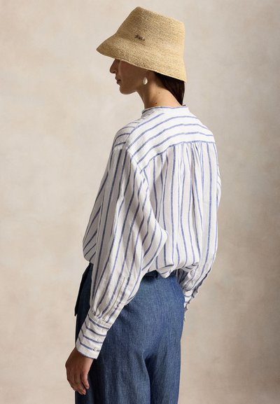 Kvinne iført en beige vevd bøttehatt, hvit bluse med blå vertikale striper og høytlivs blå bukser, sett fra siden mot en nøytral bakgrunn.