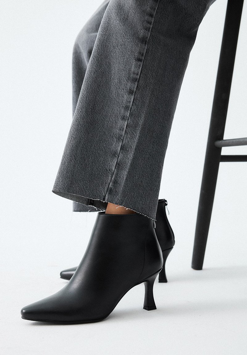 Botines de cuero negro con punta afilada y un tacón corto y delgado. Llevados con unos jeans grises de bordes deshilachados, descansando sobre una silla negra.