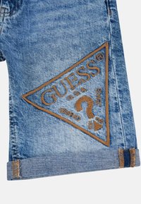 Shorts en denim bleu clair avec un ourlet replié, arborant un grand logo triangulaire brodé marron et des détails textuels d'un côté.