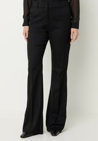 Pantaloni a palazzo neri su misura indossati con scarpe nere appuntite e una maglietta nera trasparente a maniche lunghe, mostrati dalla vita ai piedi su uno sfondo bianco.