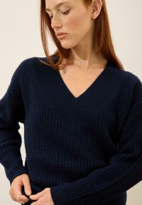 Granatowy sweter dzianinowy z głębokim dekoltem w serek, prążkowanymi mankietami oraz tkanym wzorem na strukturze materiału. Dopasowany krój z długimi rękawami.