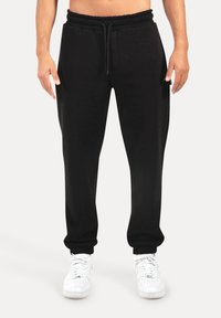 Sorte sweatpants med elastisk talje og snoretræk, med sidelommer og manchetter ved anklerne, parret med hvide sneakers.