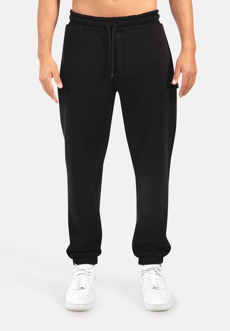 Sorte sweatpants med elastisk talje og snoretræk, med sidelommer og manchetter ved anklerne, parret med hvide sneakers.