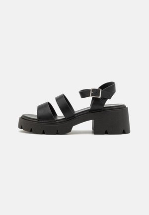Black leather sandals featuring three wide straps, an adjustable ankle buckle, and a chunky textured platform sole with a unique cutout design.