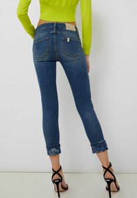 Mujer vestida con jeans azules recortados con bordados florales en los puños, un suéter corto de color verde neón y sandalias de tacón negro, de pie contra una pared blanca.