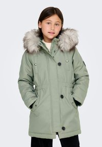 Roheline pehme vooder, millel on faux karusnahast kapuuts, lukuga sulgemine, funktsionaalsed taskud ja kitsenev vöökoht. Pehme tekstuur, nupud kaunistuseks.