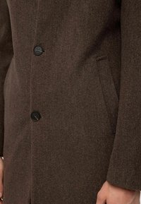 Manteau en laine marron avec tissu texturé, boutons noirs et poches latérales. Le design comprend une coupe ajustée et des lignes épurées.