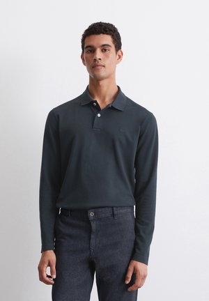 Langärmliges, dunkelblaues Poloshirt aus weichem Stoff, mit einer Drei-Knopf-Leiste und dezentem Logo auf der Brust. Kombiniert mit dunklen Jeans.